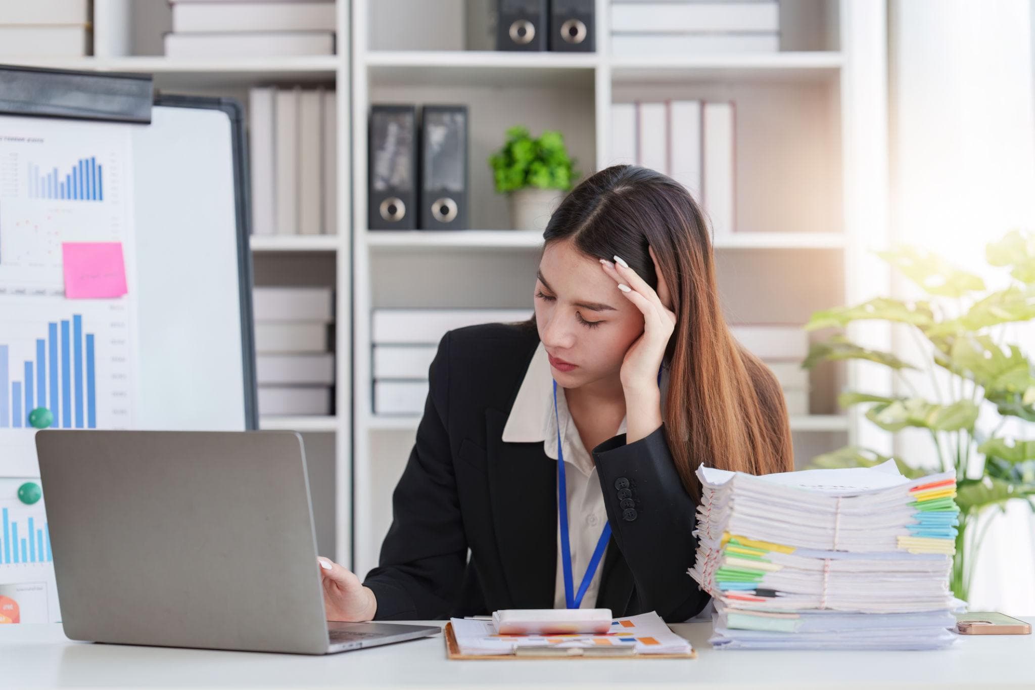 Overwhelmed businesswoman sitting at a desk with a laptop and a massive stack of documents.